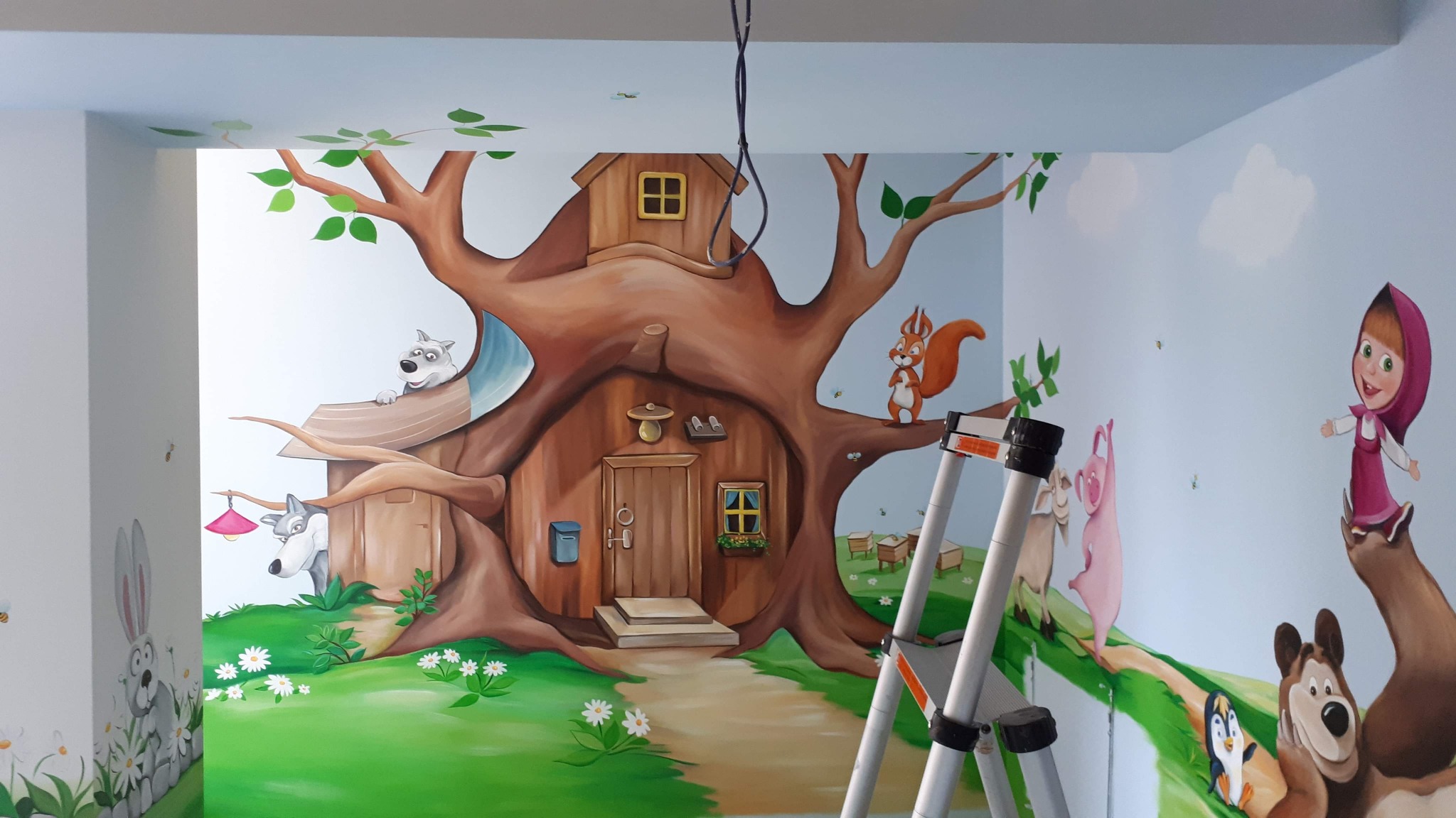 Ręcznie malowana ściana w pokoju dziecięcym przedstawiająca bajkowy las z domkiem w drzewie, postaciami zwierząt i postacią z bajki Masza i Niedźwiedź, widoczna drabina malarska.