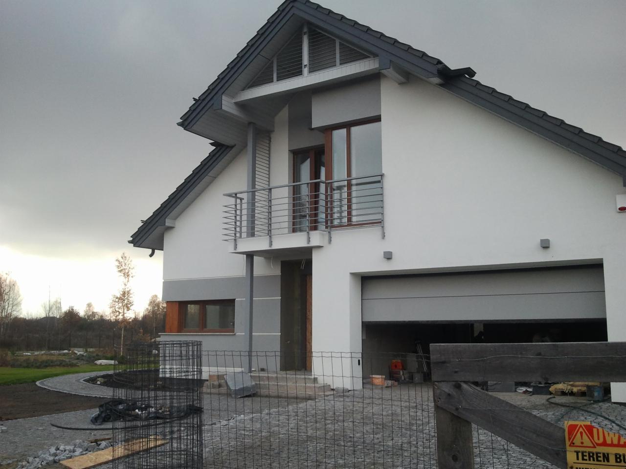 Nowoczesny dom jednorodzinny z balkonem i otwartym garażem, widoczna kostka brukowa na podjeździe i siatka zabezpieczająca teren budowy.