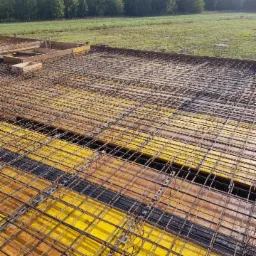 Zbrojenie płyty fundamentowej na żółtym szalunku, widok z góry na siatkę zbrojeniową przed betonowaniem, w tle zielone pole i drzewa.