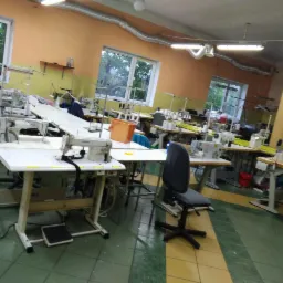 Wnętrze szwalni z wieloma stanowiskami pracy wyposażonymi w maszyny do szycia JUKI, widoczne okna i oświetlenie sufitowe.