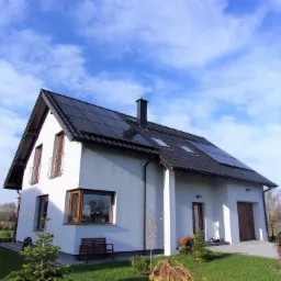 Nowoczesny dom jednorodzinny z białymi ścianami, ciemną dachówką i panelami słonecznymi, widoczny z perspektywy ogrodu z młodymi drzewkami iglastymi i samochodem zaparkowanym obok garażu.