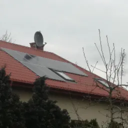 Panele słoneczne na czerwonym dachu domu jednorodzinnego z anteną satelitarną na kominie, widoczne również okna dachowe.