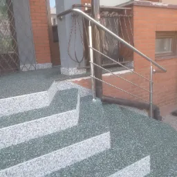 Zewnętrzne schody wejściowe wykończone żywicą epoksydową z widoczną fakturą kamyczkową, zabezpieczone metalową balustradą i rynną spustową.