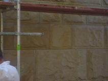 Piaskowcowa elewacja w trakcie czyszczenia pod rusztowaniem, widoczne ślady impregnacji i płomieniowania, fragment sylwetki pracownika w białym kombinezonie.