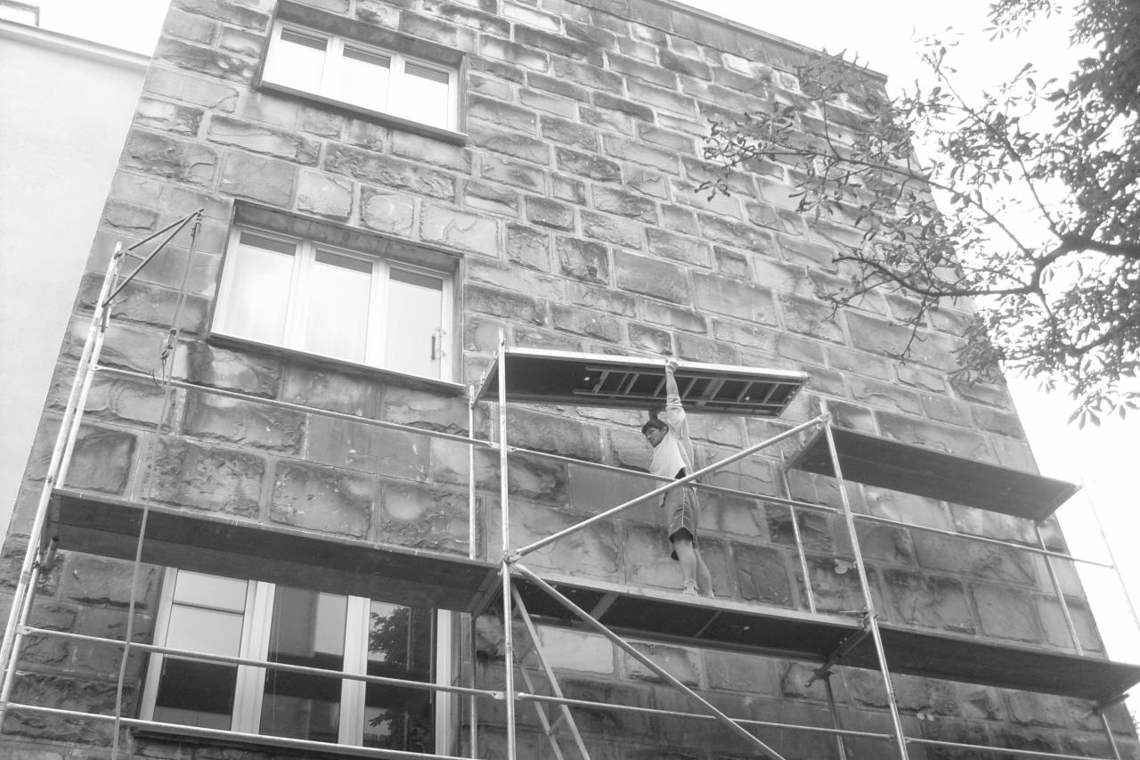 Czarno-białe zdjęcie elewacji budynku z kamienia, na której ustawione jest rusztowanie, a na nim mężczyzna przenosi aluminiową drabinę.