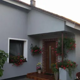 Szary dom jednorodzinny z brązowym dachem, oknami z białymi ramami i dekoracjami kwiatowymi przy wejściu i pod oknami.