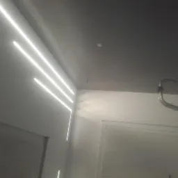 Nowoczesne oświetlenie LED w postaci trzech równoległych linii na ścianie, z ciemnym sufitem i fragmentem drabiny malarskiej w rogu