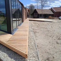 Nowoczesny taras z desek modrzewiowych, przylegający do budynku z przeszkloną ścianą, obok Muzeum Garncarstwa, z widoczną podsypką z białego kamienia i przygotowanym terenem pod dalsze prace.