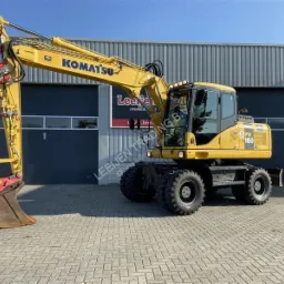 Żółta koparka kołowa Komatsu PW160 zaparkowana na brukowanym placu przed szarym budynkiem z zamkniętą bramą garażową.