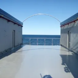 Biało-szara, gładka powierzchnia tarasu z widokiem na morze, ograniczonego szklaną balustradą i budynkami po bokach, połączonymi metalowym łukiem.