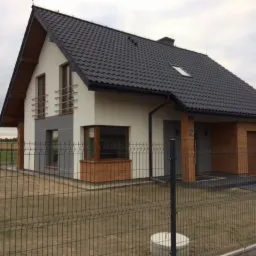 Nowoczesny dom murowany z czarną dachówką, drewnianymi elementami elewacji i metalowym ogrodzeniem panelowym na działce z widokiem na pole.