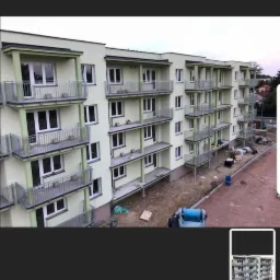 Wielopiętrowy budynek mieszkalny w trakcie budowy, z widocznymi balkonami z metalowymi barierkami, teren budowy z nieutwardzonym podłożem i sprzętem budowlanym.