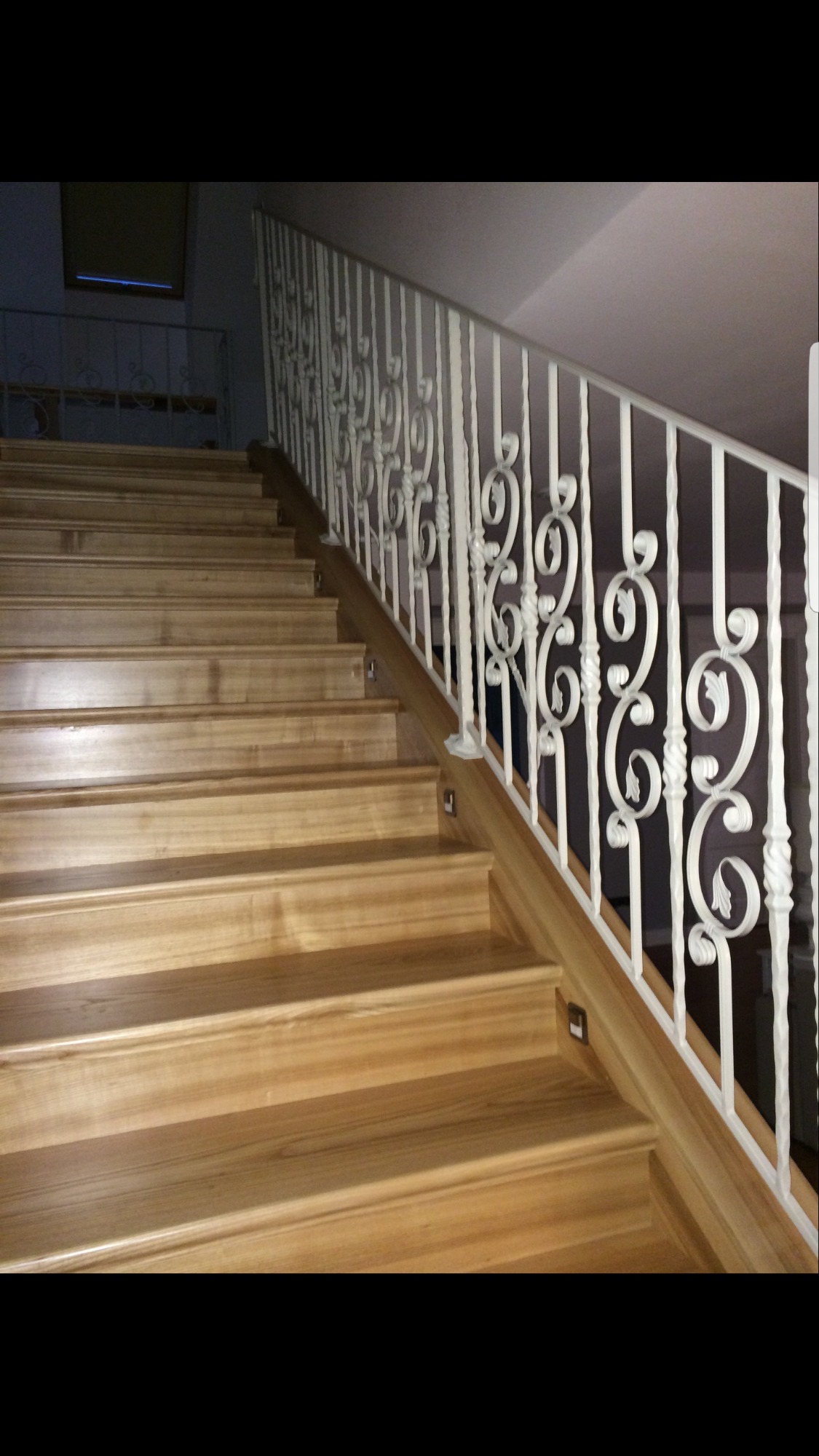 Jasne drewniane schody z białą, ozdobną metalową barierką i subtelnym oświetleniem w stopniach.