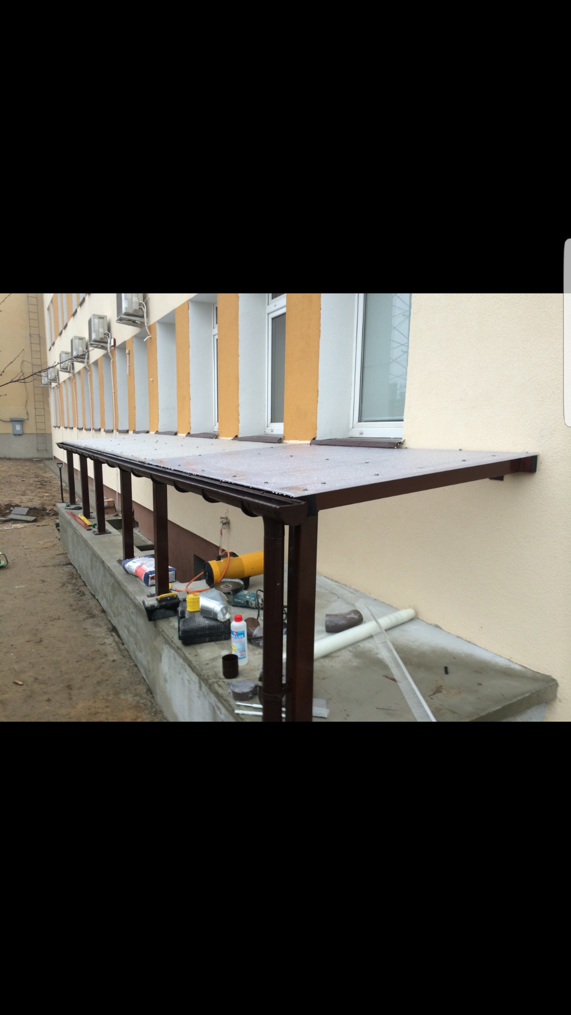 Zadaszenie nad wejściem do budynku, konstrukcja metalowa w kolorze brązowym, przezroczysty dach z poliwęglanu, widoczne narzędzia i materiały budowlane na betonowym podłożu.