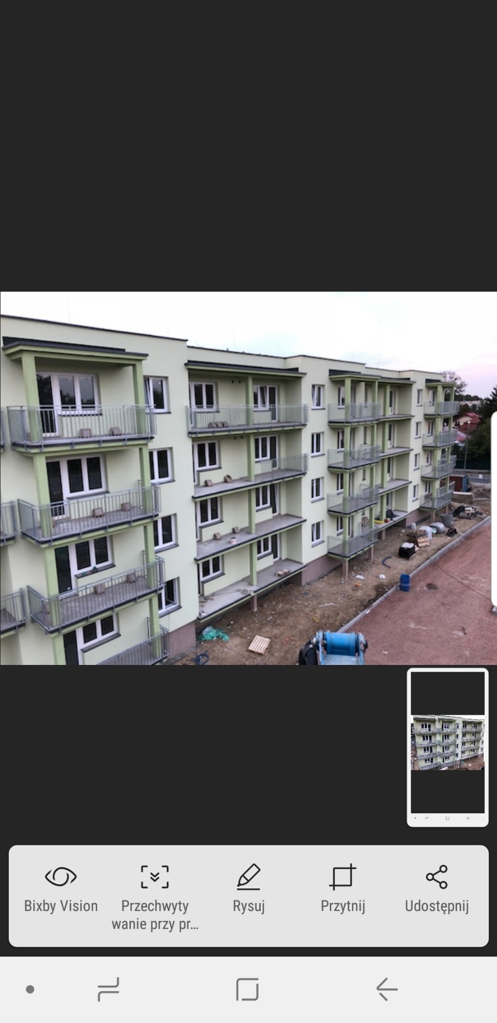 Wielopiętrowy budynek mieszkalny w trakcie budowy, z widocznymi balkonami z metalowymi barierkami, teren budowy z nieutwardzonym podłożem i sprzętem budowlanym.