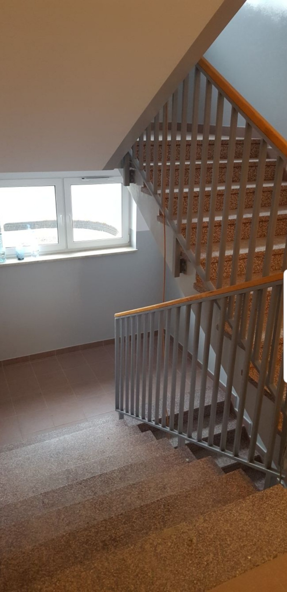 Klatka schodowa z szarymi barierkami z drewnianym uchwytem, widok z dołu na górę, okno z butelkami w tle.