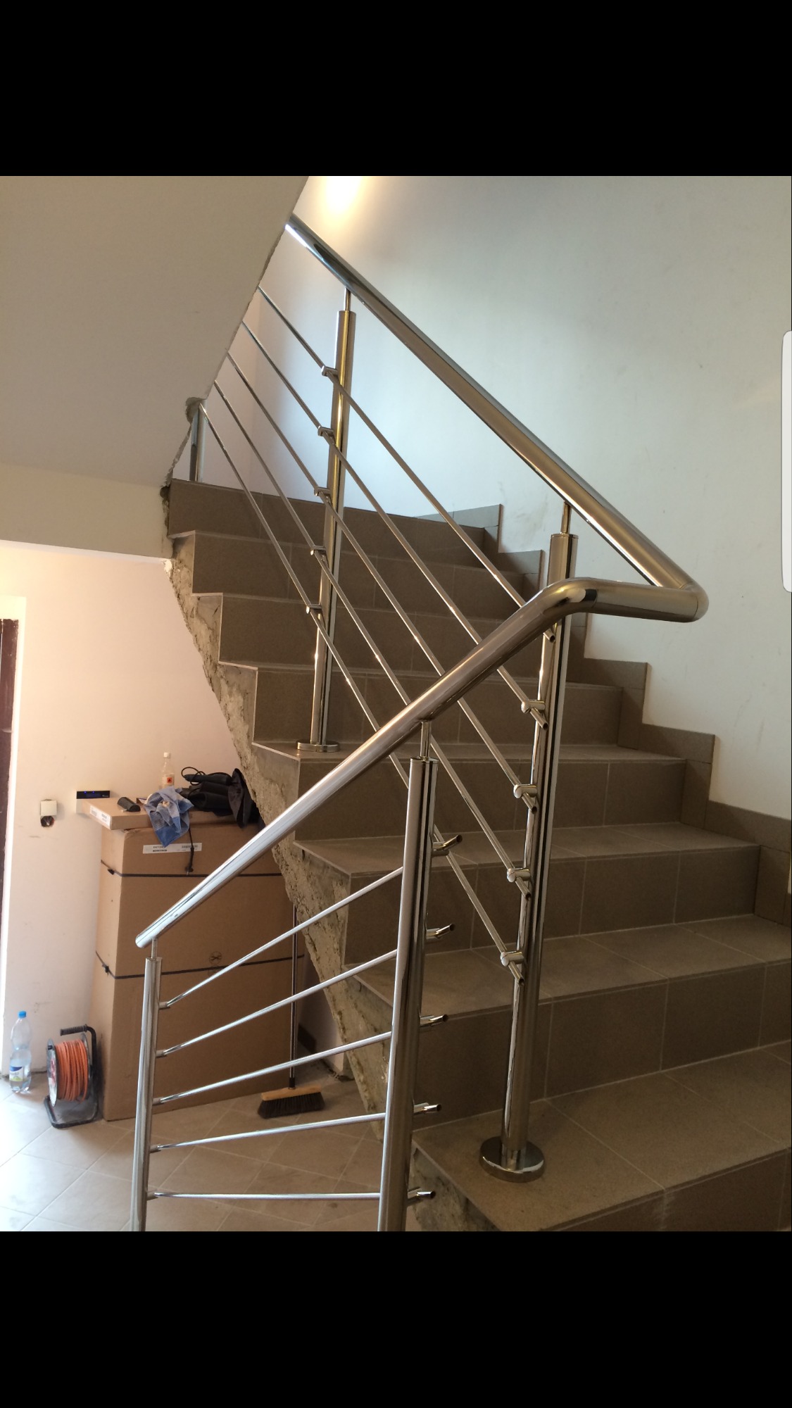 Schody wykończone brązowymi płytkami z zamontowaną, nowoczesną, metalową barierką z poziomymi prętami. Widoczne elementy wykończeniowe i narzędzia na kartonach w tle.