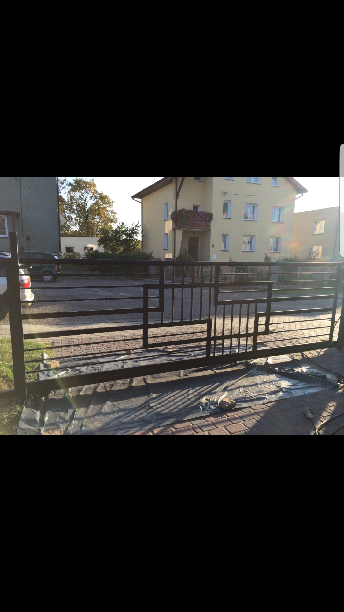 Czarna, metalowa brama przesuwna o geometrycznym wzorze, częściowo osłonięta folią malarską, na tle kostki brukowej i budynku mieszkalnego w słoneczny dzień.