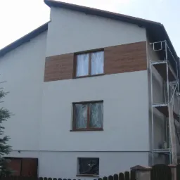 Dwukondygnacyjny dom po ociepleniu, z drewnianą wstawką na elewacji, okna zabezpieczone folią, rusztowanie z boku budynku, ogrodzenie i drzewo na pierwszym planie.