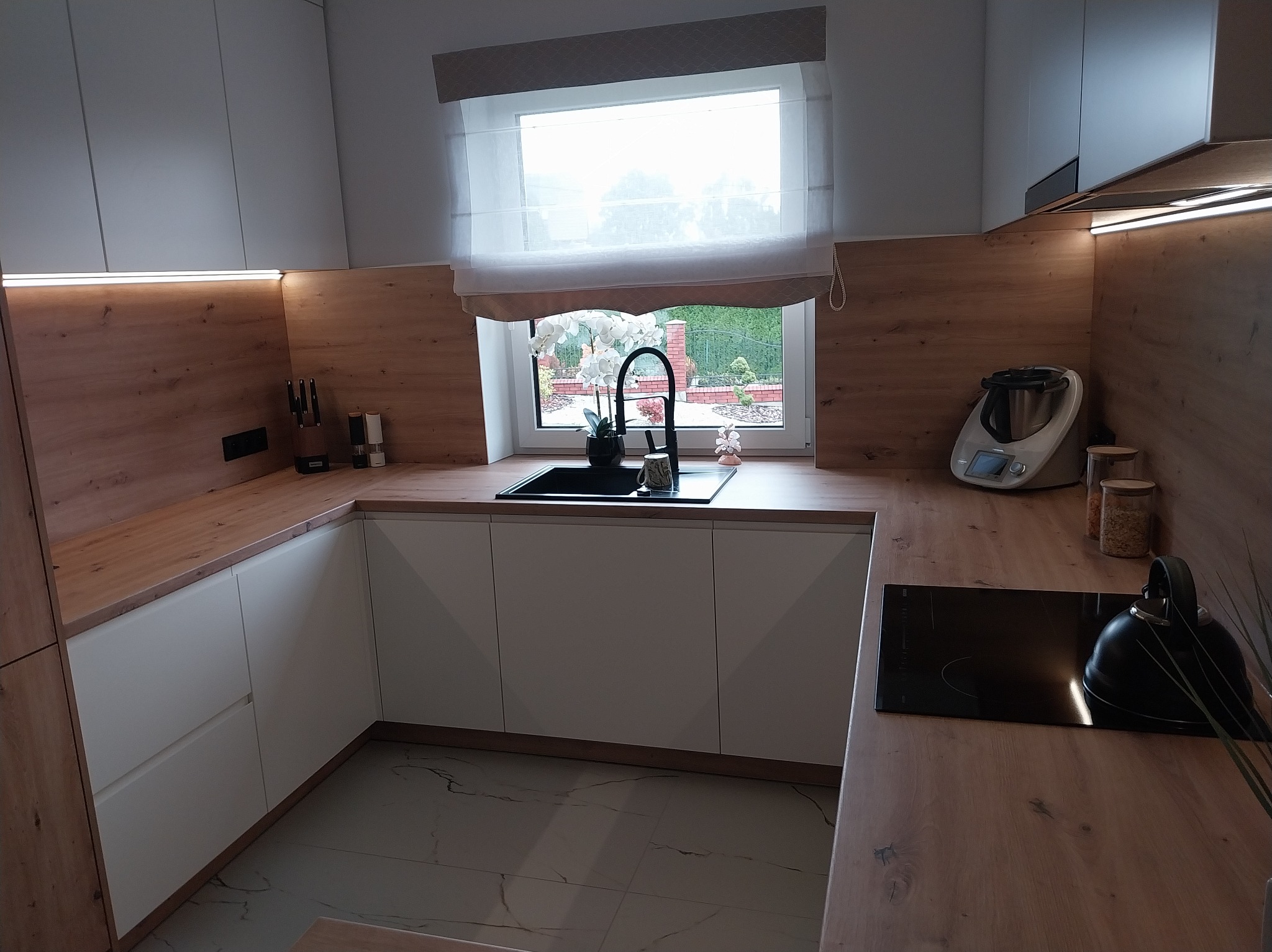 Nowoczesna kuchnia z białymi szafkami i drewnianymi blatami. Widok na okno z roletą rzymską i ogród. Czarny zlew i płyta indukcyjna. Minimalistyczny design.