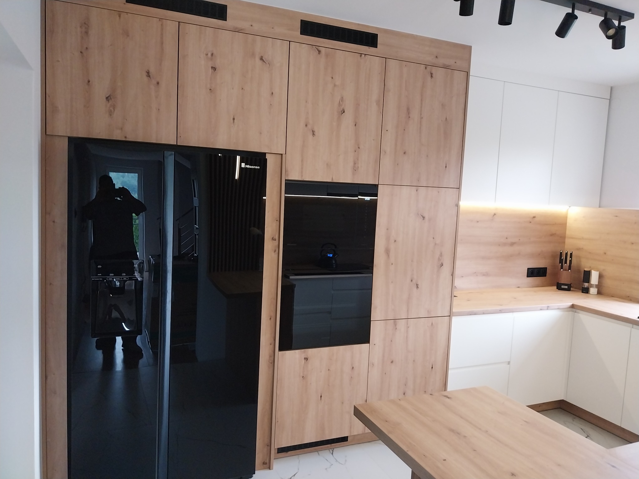 Nowoczesna kuchnia z zabudową w odcieniach drewna i czerni, z widocznym sprzętem AGD i oświetleniem punktowym. Minimalistyczny design z elementami dekoracyjnymi.