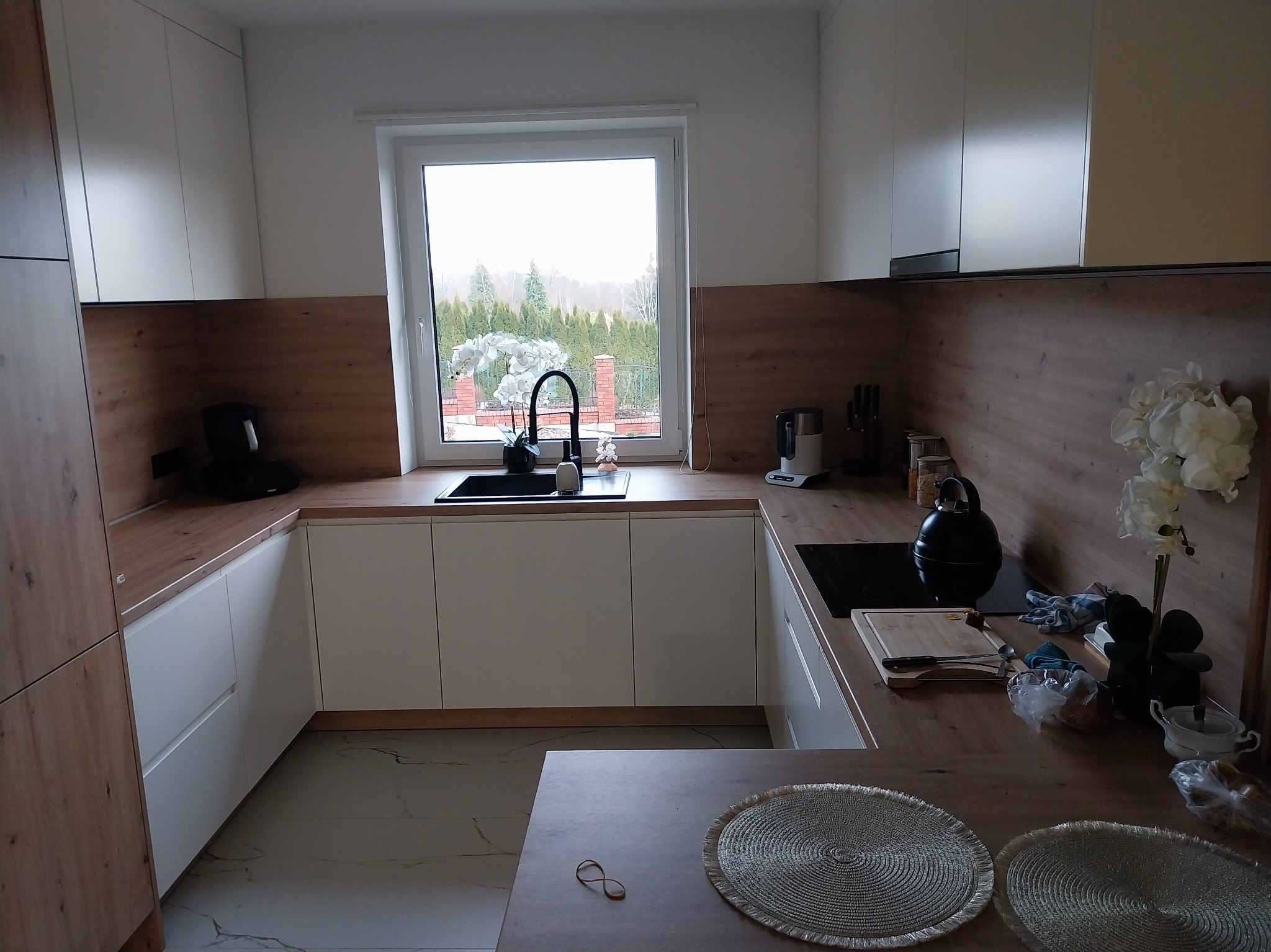 Nowoczesna kuchnia w bieli i drewnie z widokiem na ogród przez okno. Minimalistyczny design, czarne akcenty, marmurowe płytki podłogowe i dekoracyjne podkładki na blacie.