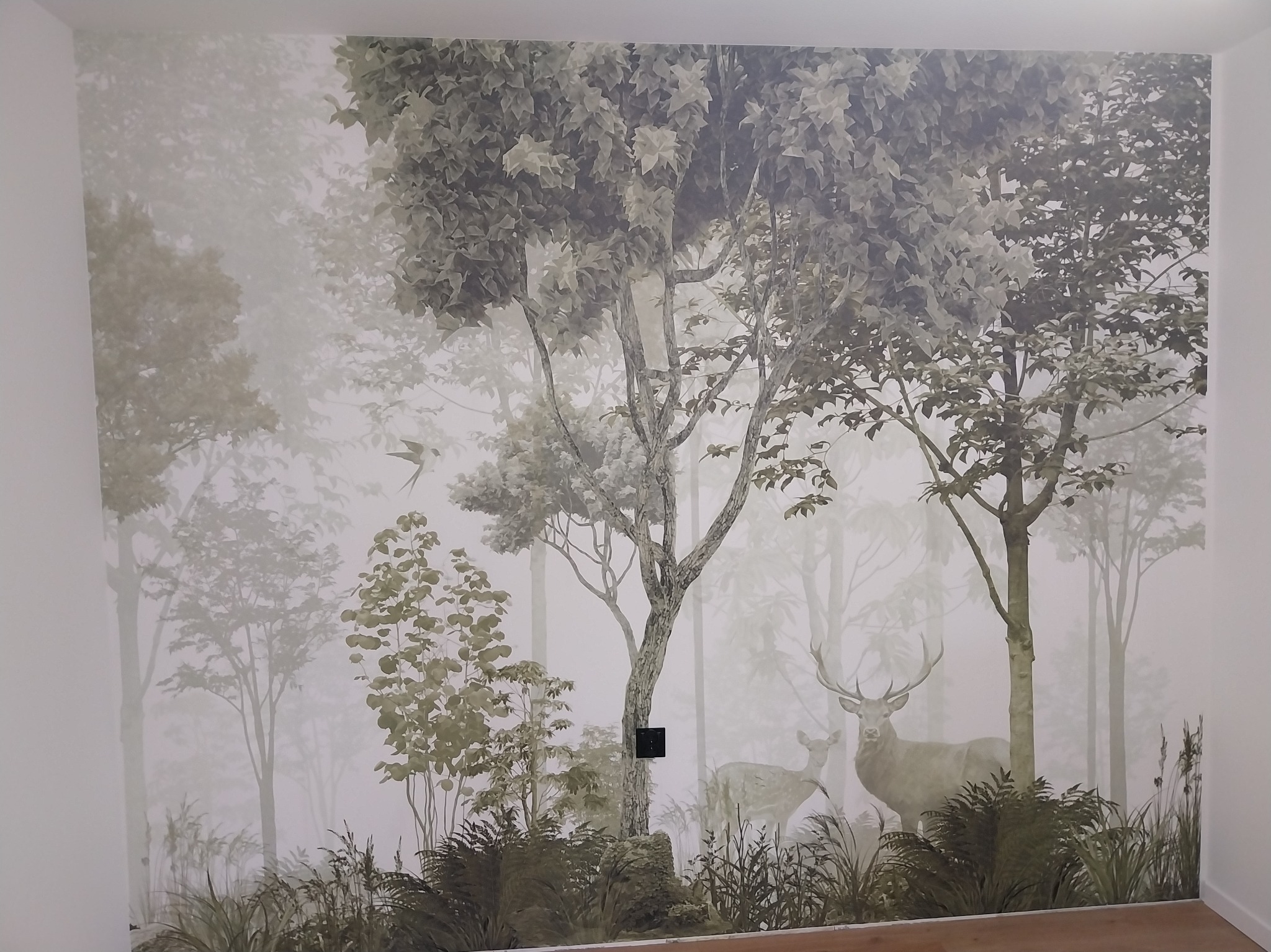 Fototapeta w pokoju z motywem leśnym: drzewa, jelenie i mgła, tworząca unikalny, naturalistyczny klimat wnętrza. Widoczny włącznik na ścianie.
