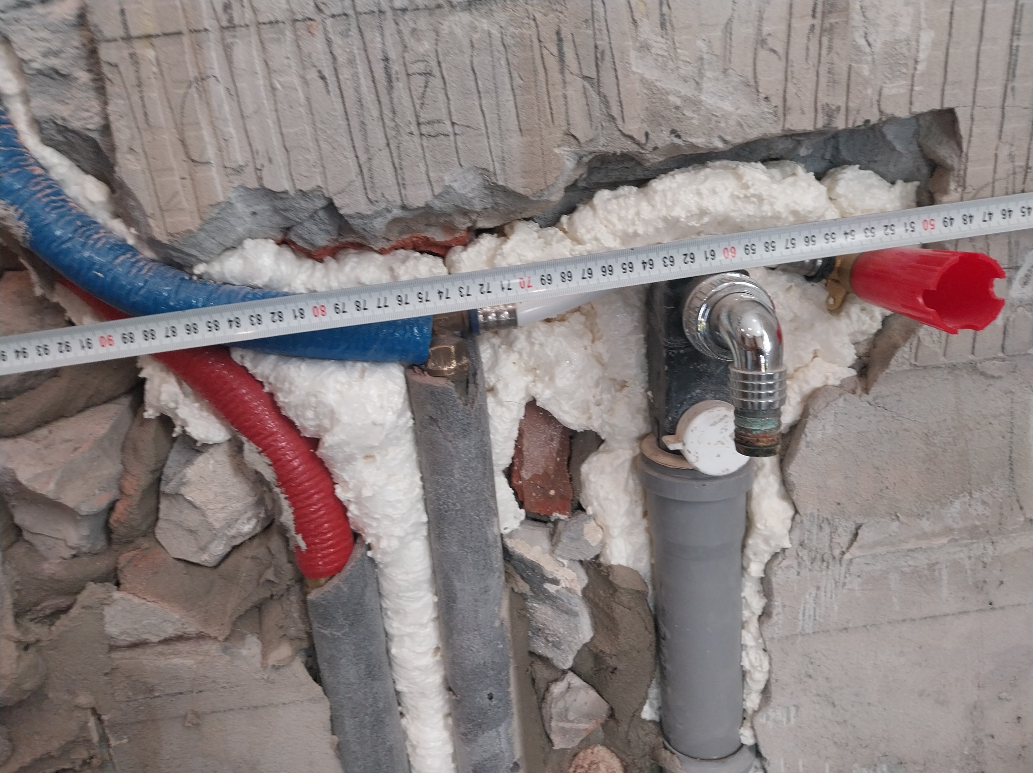 Instalacja wodno-kanalizacyjna w trakcie montażu, rury w izolacji piankowej, pomiar odległości miarką na tle surowej ściany z widocznymi śladami po kuciu.