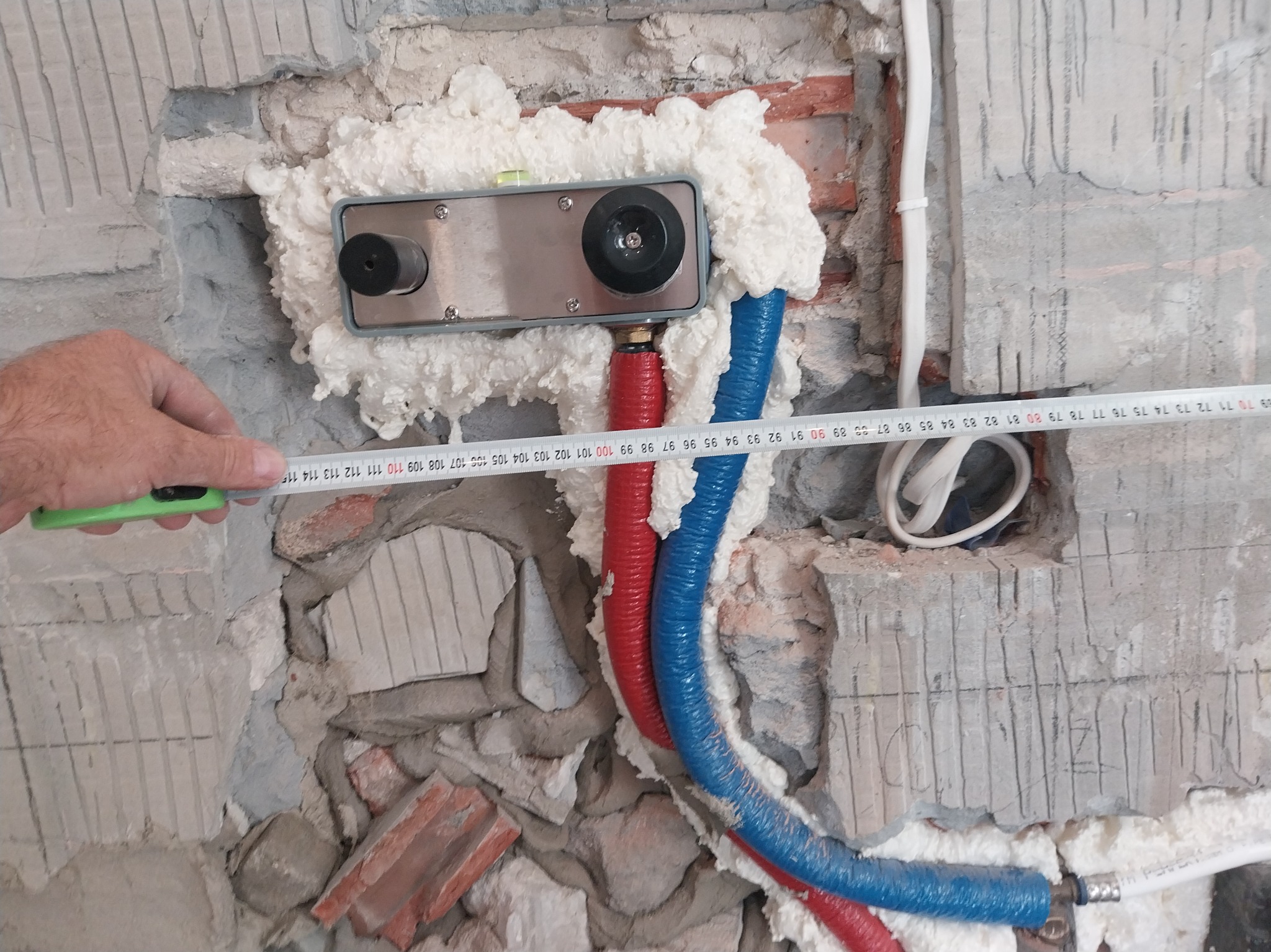 Montaż podtynkowego zestawu prysznicowego z pianką montażową i rurami PEX w ścianie. Mierzenie odległości miarką. Widoczne ślady po kuciu w ścianie.