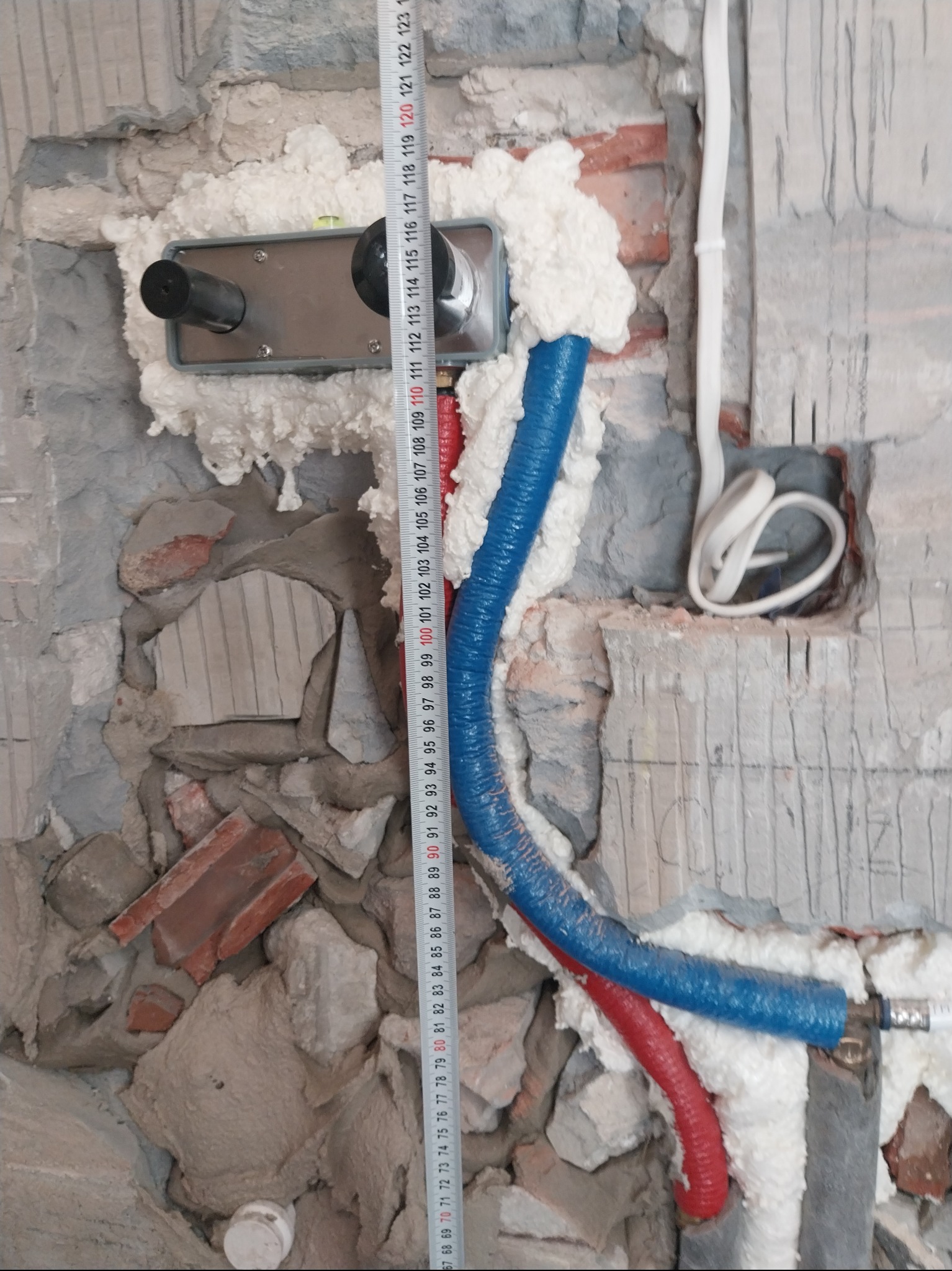Montaż instalacji wodnej w łazience: rury (czerwona i niebieska) zabezpieczone pianką montażową, widoczna miarka i fragment ściany z instalacją elektryczną.