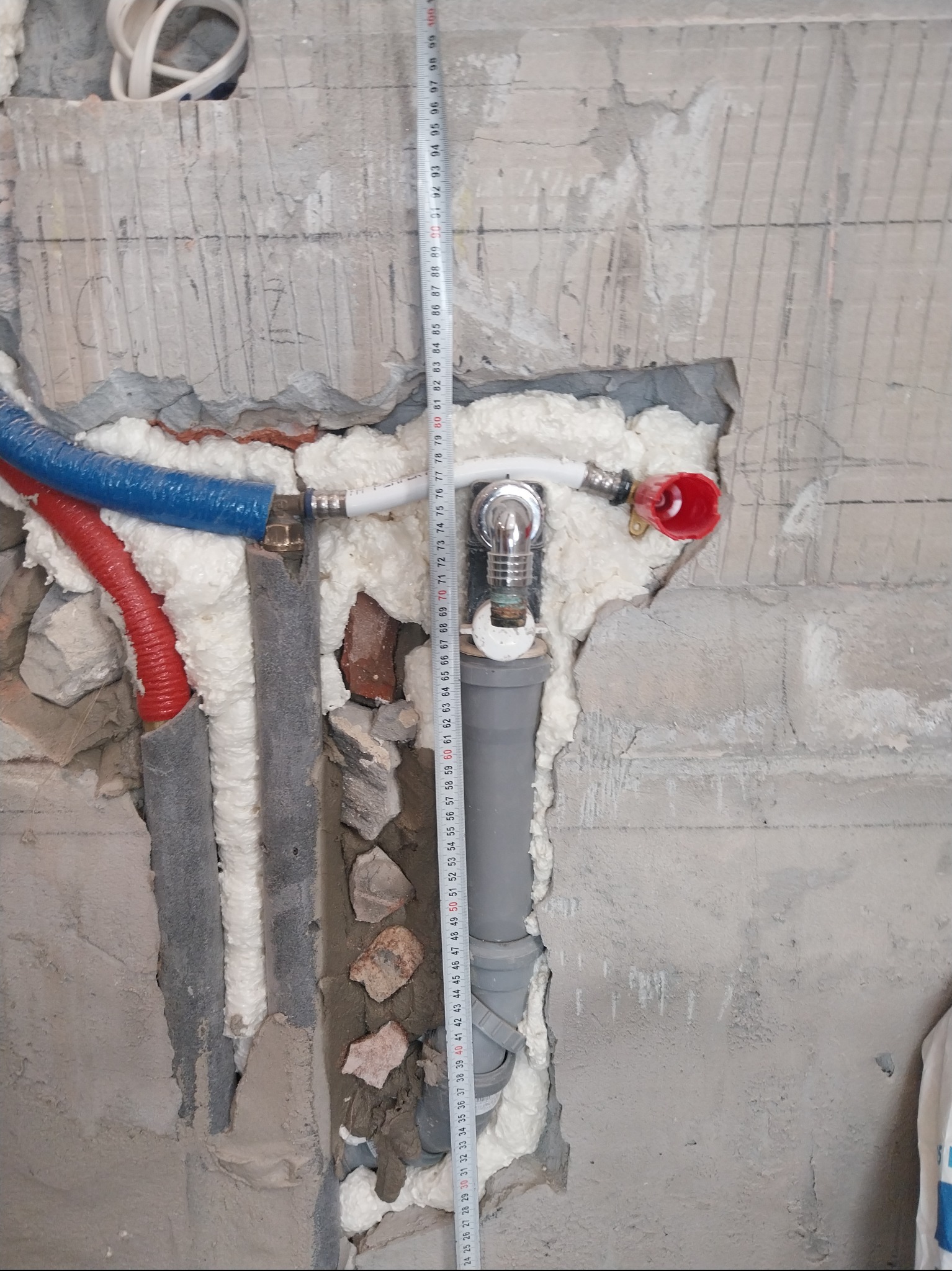 Instalacja wodno-kanalizacyjna w surowym stanie, rury zabezpieczone pianką montażową, widoczna miarka. Prace instalacyjne w trakcie remontu łazienki.