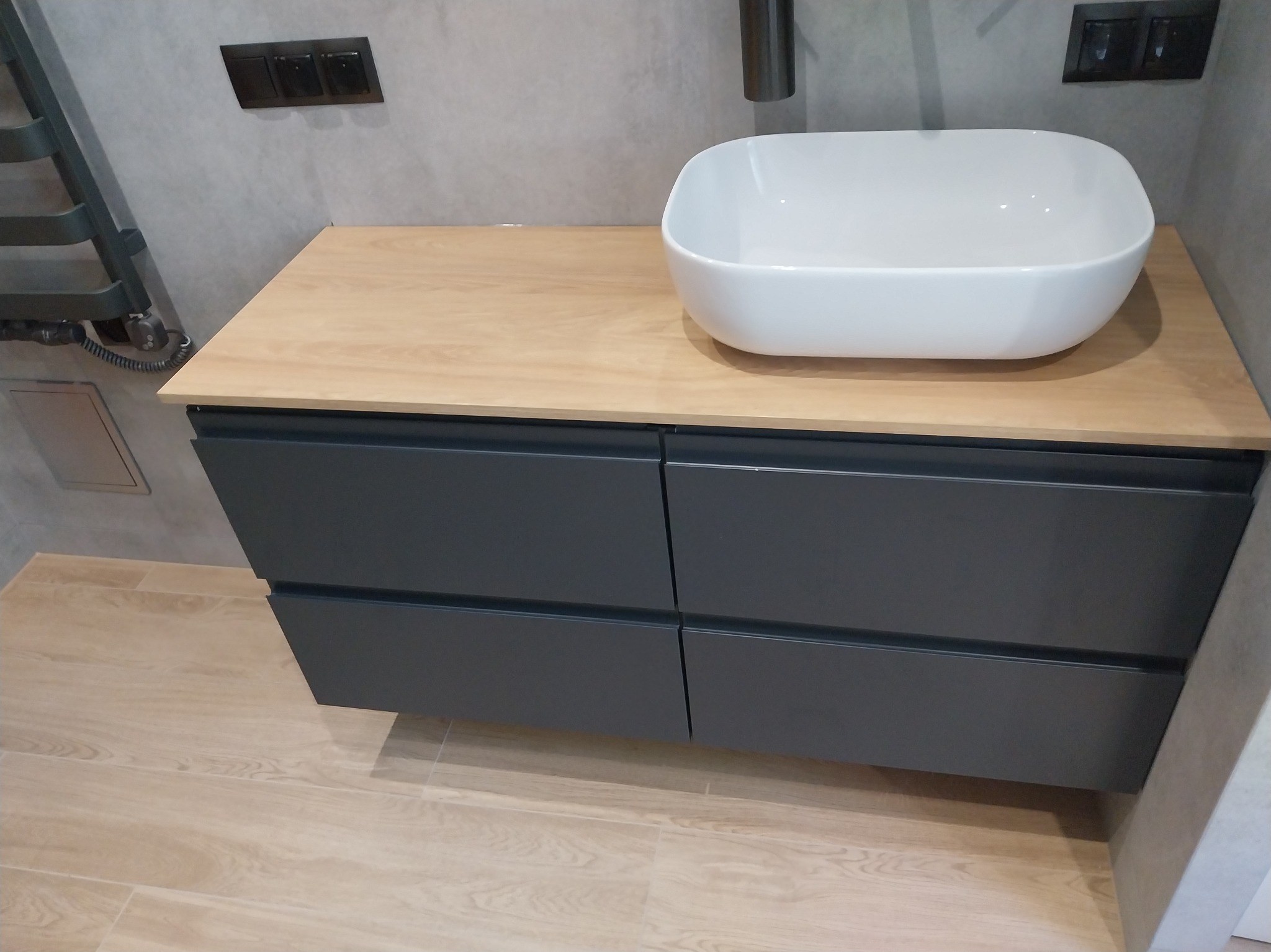 Nowoczesna łazienka: biała umywalka nablatowa na szafce z szufladami w kolorze grafitowym, drewniany blat, płytki podłogowe imitujące drewno, minimalistyczne wykończenie.