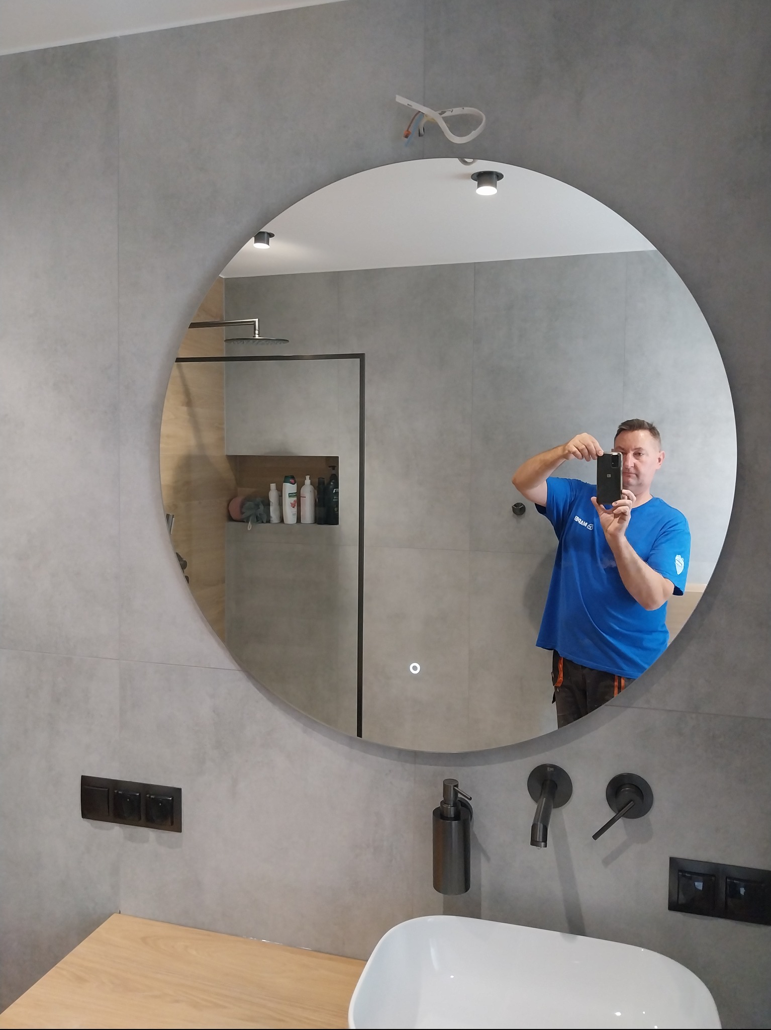 Szara łazienka z okrągłym lustrem, w którym odbija się mężczyzna robiący zdjęcie. Widoczne czarne baterie, umywalka i wnęka prysznicowa z kosmetykami.