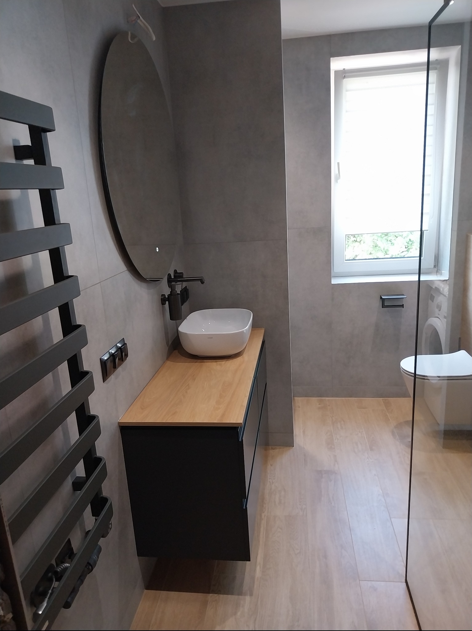 Nowoczesna łazienka z szarą glazurą, drewnianym blatem pod umywalką i okrągłym lustrem z podświetleniem. Widoczny grzejnik drabinkowy i kabina prysznicowa ze szkła.