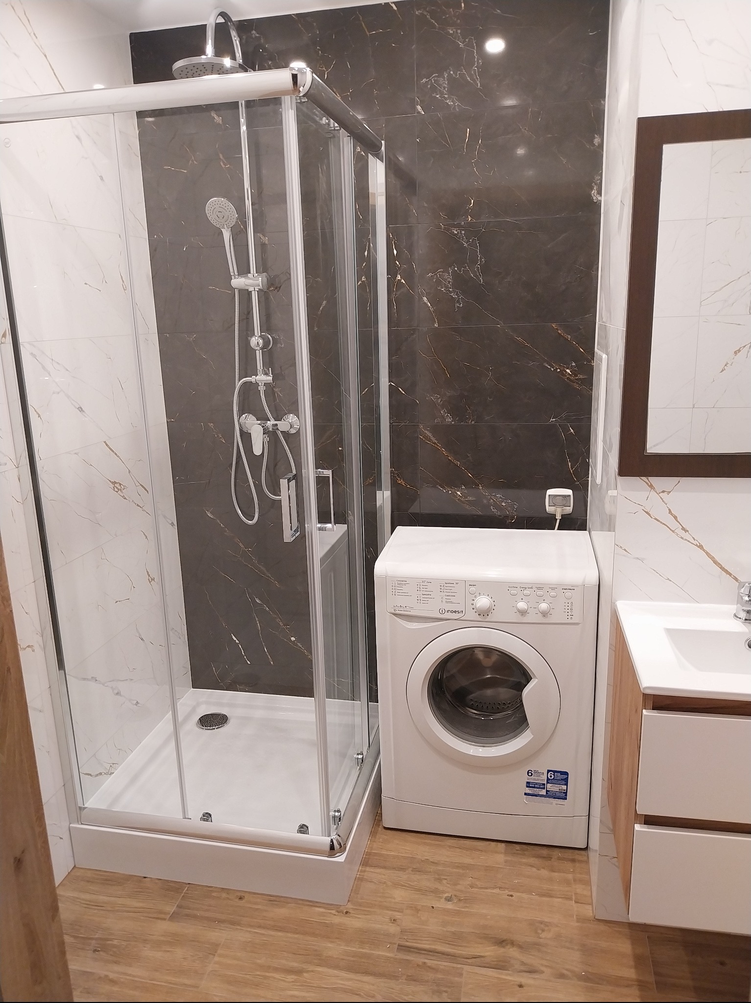 Nowoczesna łazienka po remoncie: kabina prysznicowa z deszczownicą, pralka Indesit i umywalka z szafką na tle płytek imitujących marmur.
