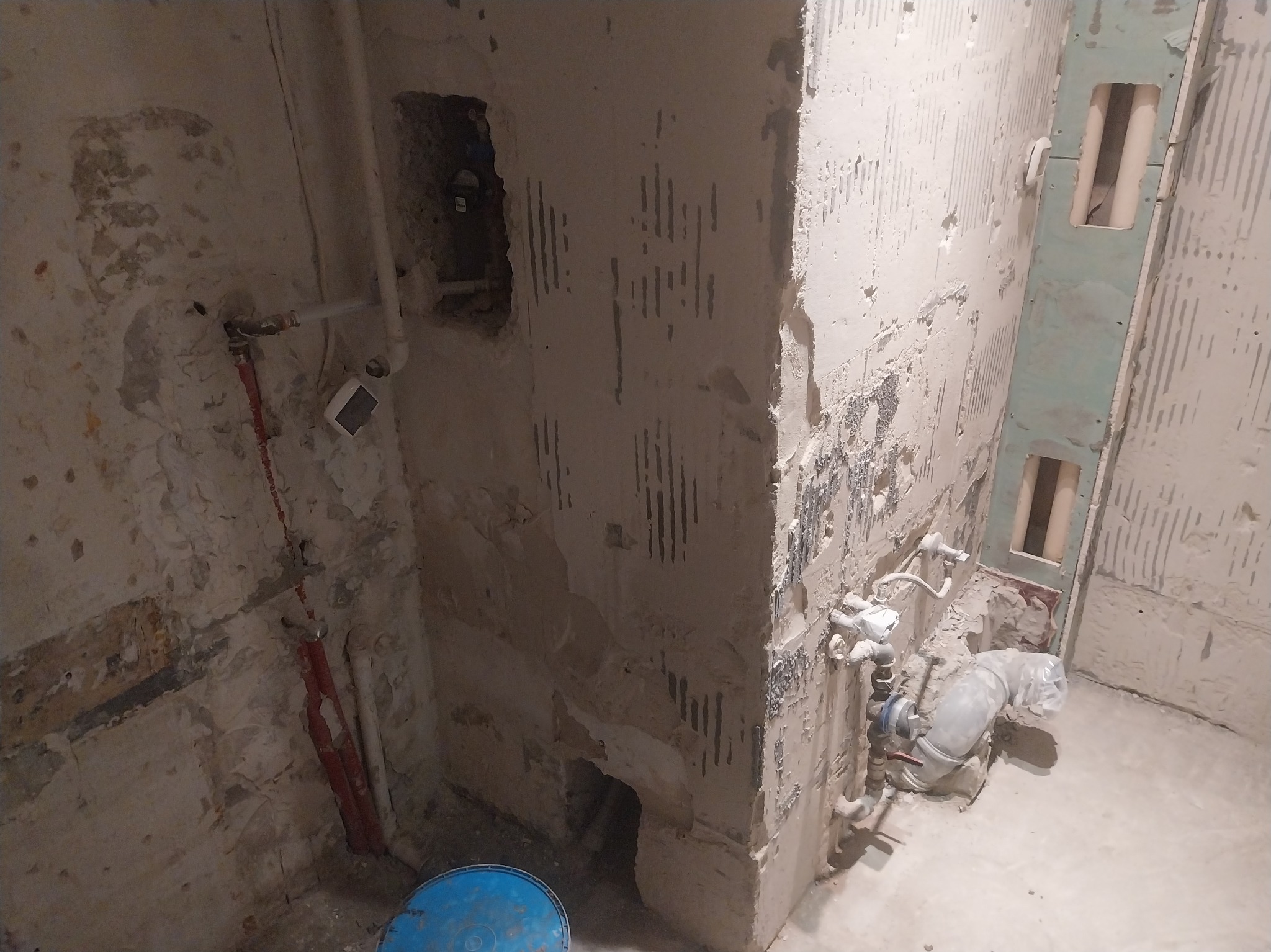 Wnętrze łazienki w trakcie remontu: ściany przygotowane pod płytki, widoczne instalacje wodne i kanalizacyjne, fragment niebieskiego wiadra na podłodze. Surowy stan przed wykończeniem.