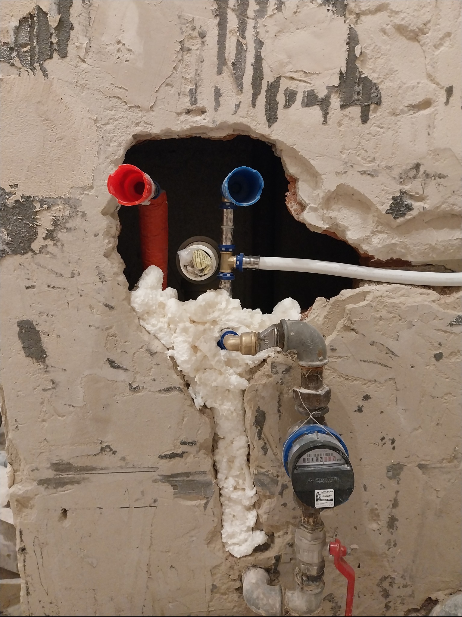 Instalacja wodna w surowej ścianie z pianką montażową, widoczne rury z kolorowymi końcówkami i licznik wody. Prace hydrauliczne w trakcie remontu.
