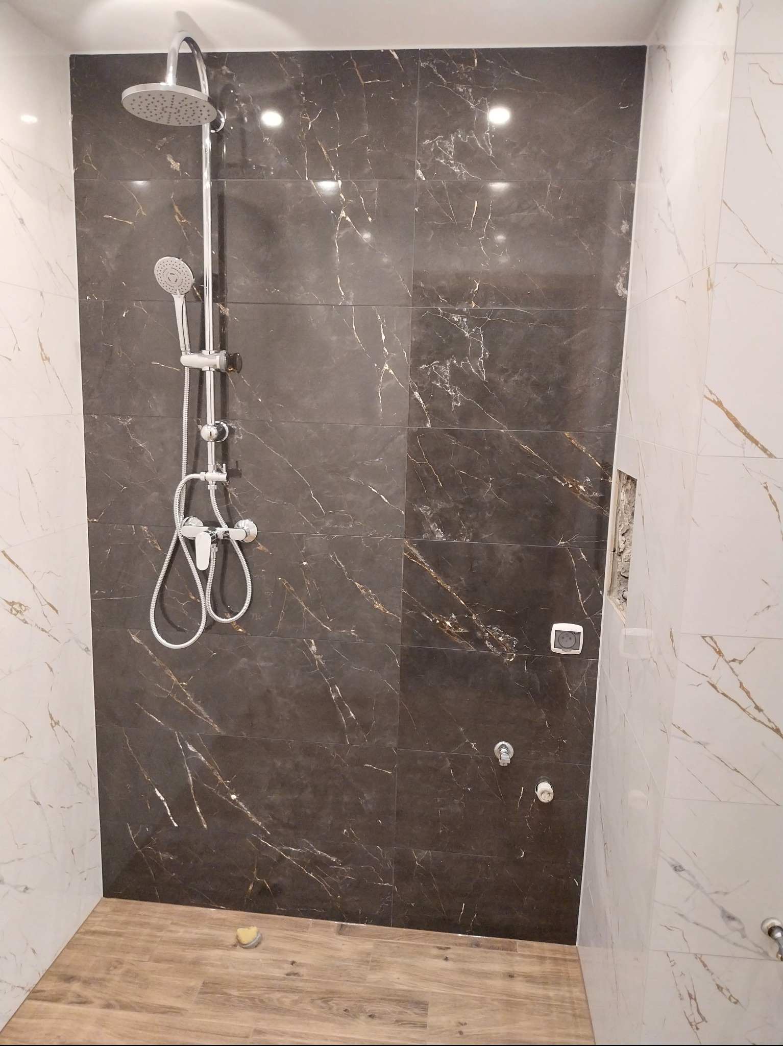 Wykończona kabina prysznicowa z ciemnymi płytkami imitującymi marmur, jasnymi płytkami na bocznych ścianach i drewnopodobną podłogą. Widoczny zestaw prysznicowy i otwór na gniazdko.