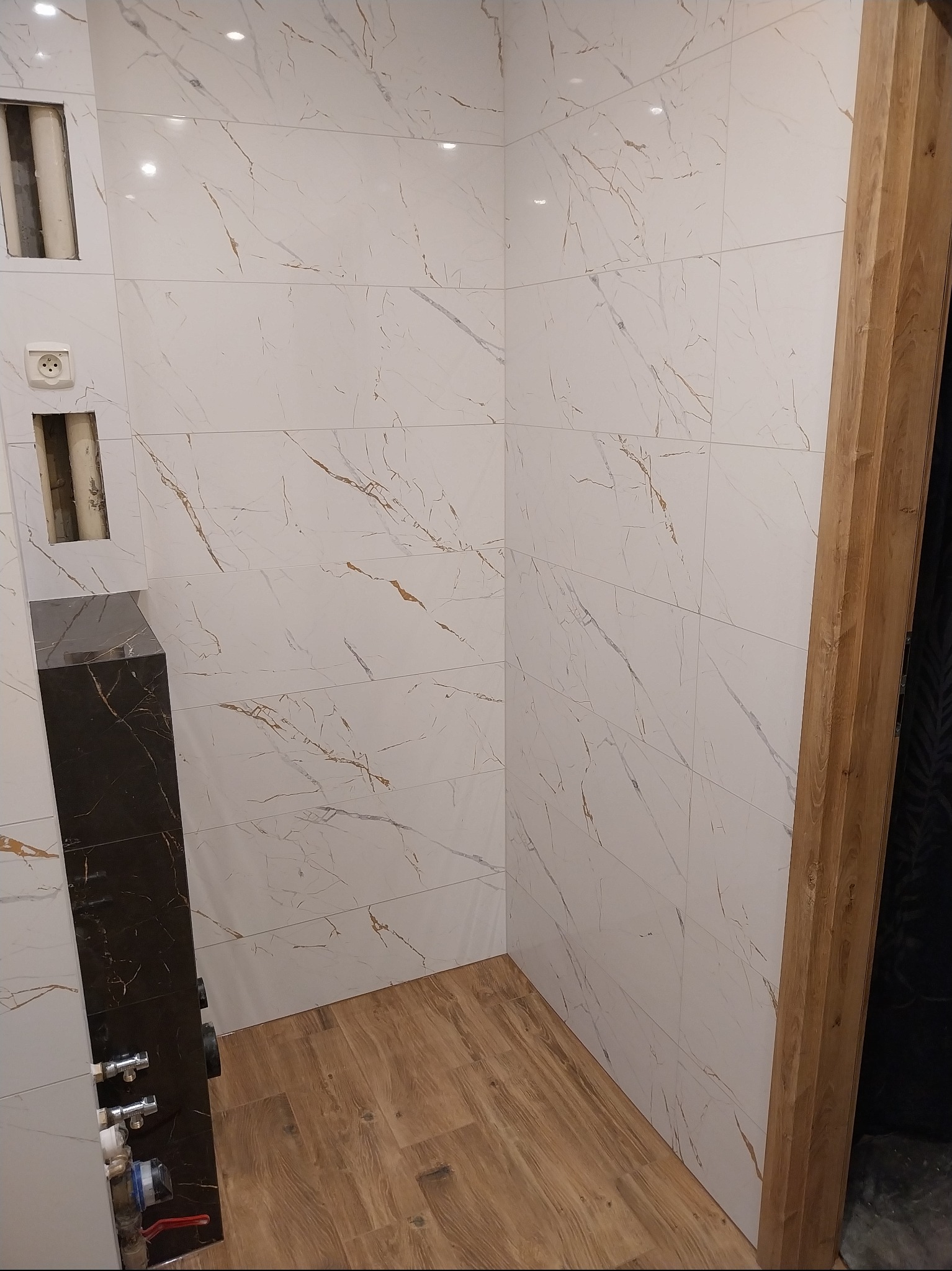 Wykończona łazienka z białymi płytkami imitującymi marmur z delikatnym złotym żyłowaniem, drewnopodobna podłoga i fragment czarnej obudowy. Widoczne instalacje.