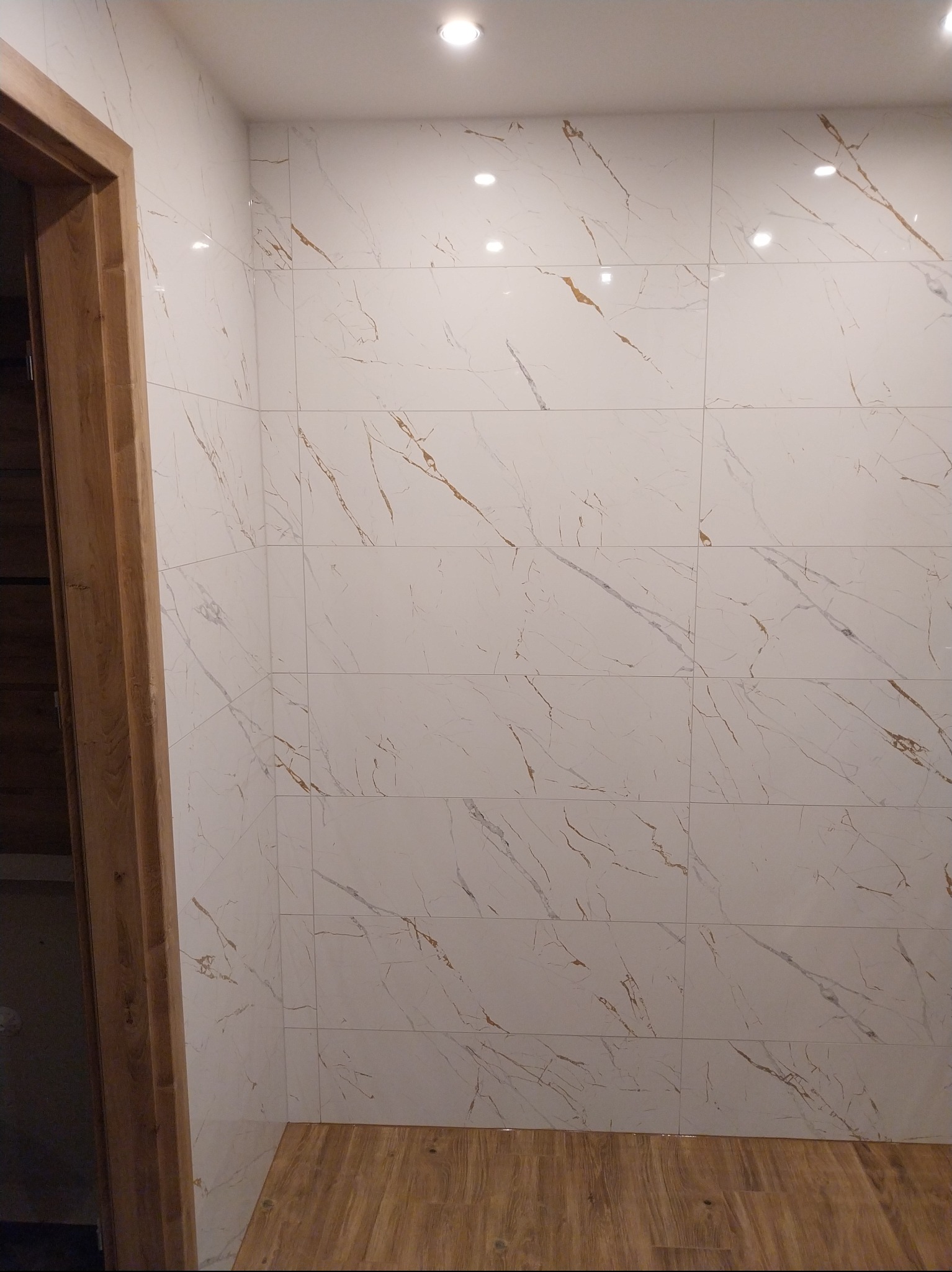 Wykończona łazienka z białymi płytkami imitującymi marmur z brązowymi żyłkami, drewnopodobna podłoga i framuga drzwi. Nowoczesny design.