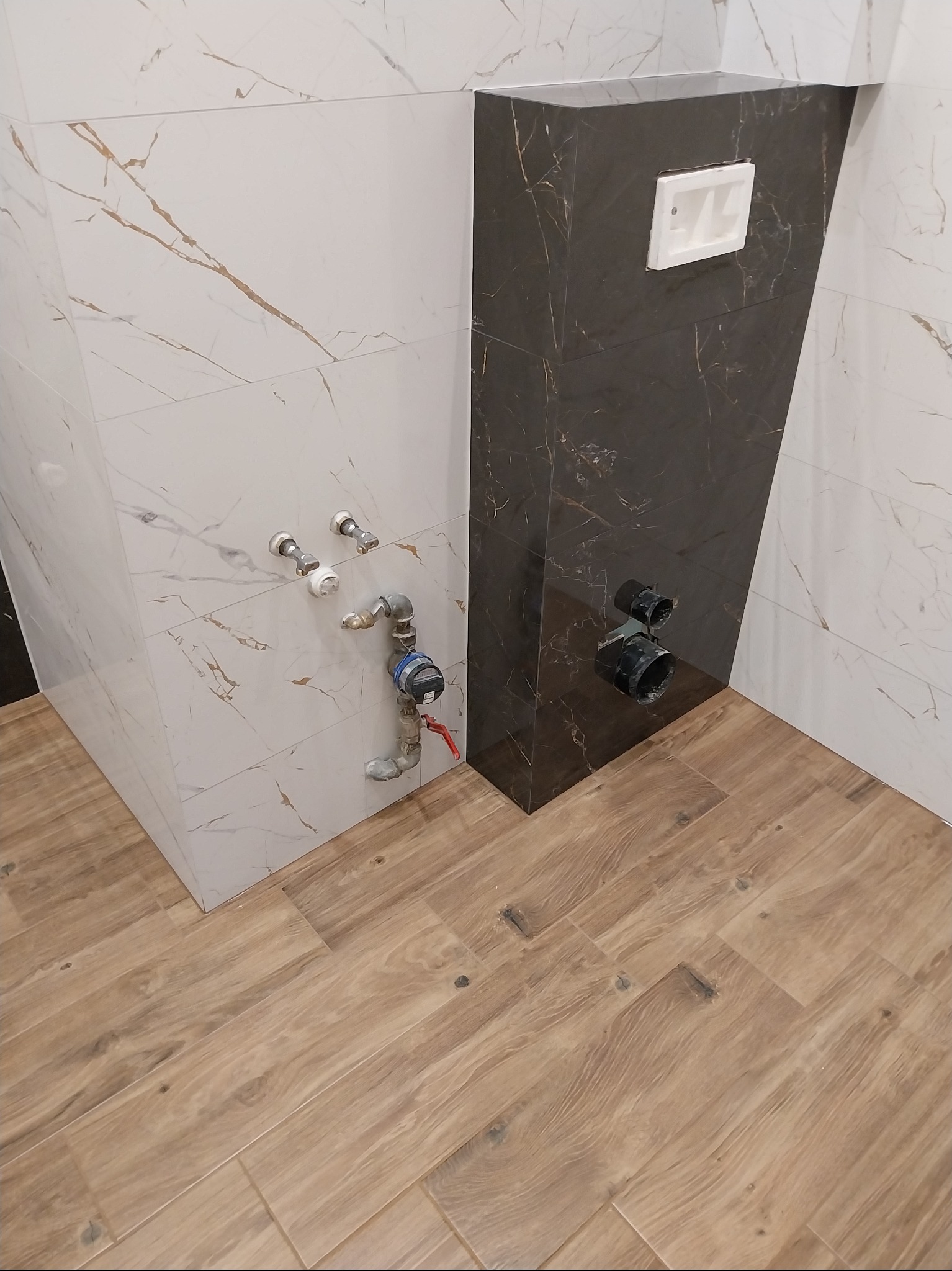 Wykończona łazienka z brązowymi panelami podłogowymi imitującymi drewno i fragmentem ściany z białych i czarnych płytek imitujących marmur z instalacją wodną.