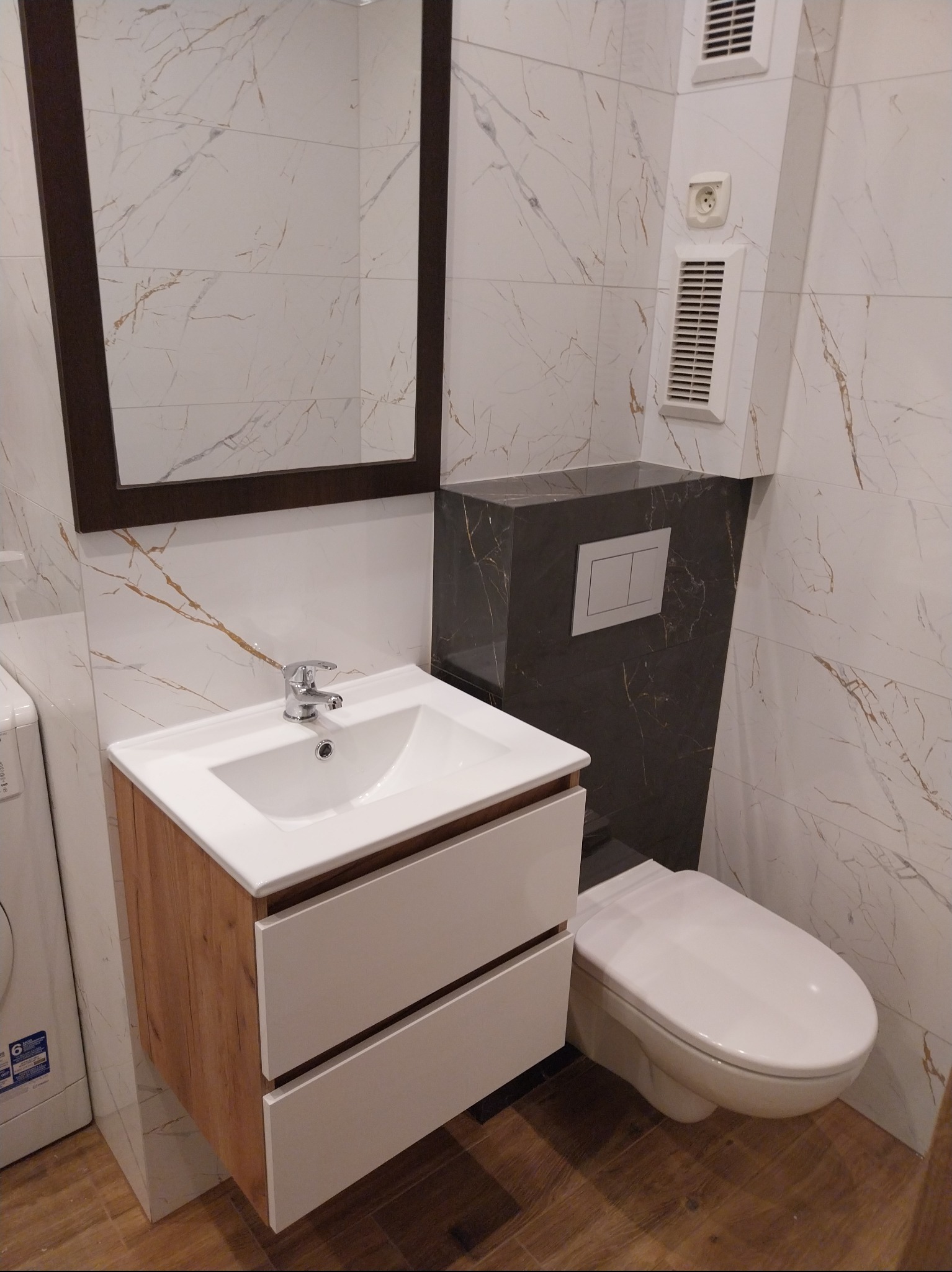 Nowoczesna łazienka z umywalką na szafce z drewnianym frontem, lustrem w ciemnej ramie i podwieszaną toaletą. Marmurowe płytki na ścianach i podłodze.