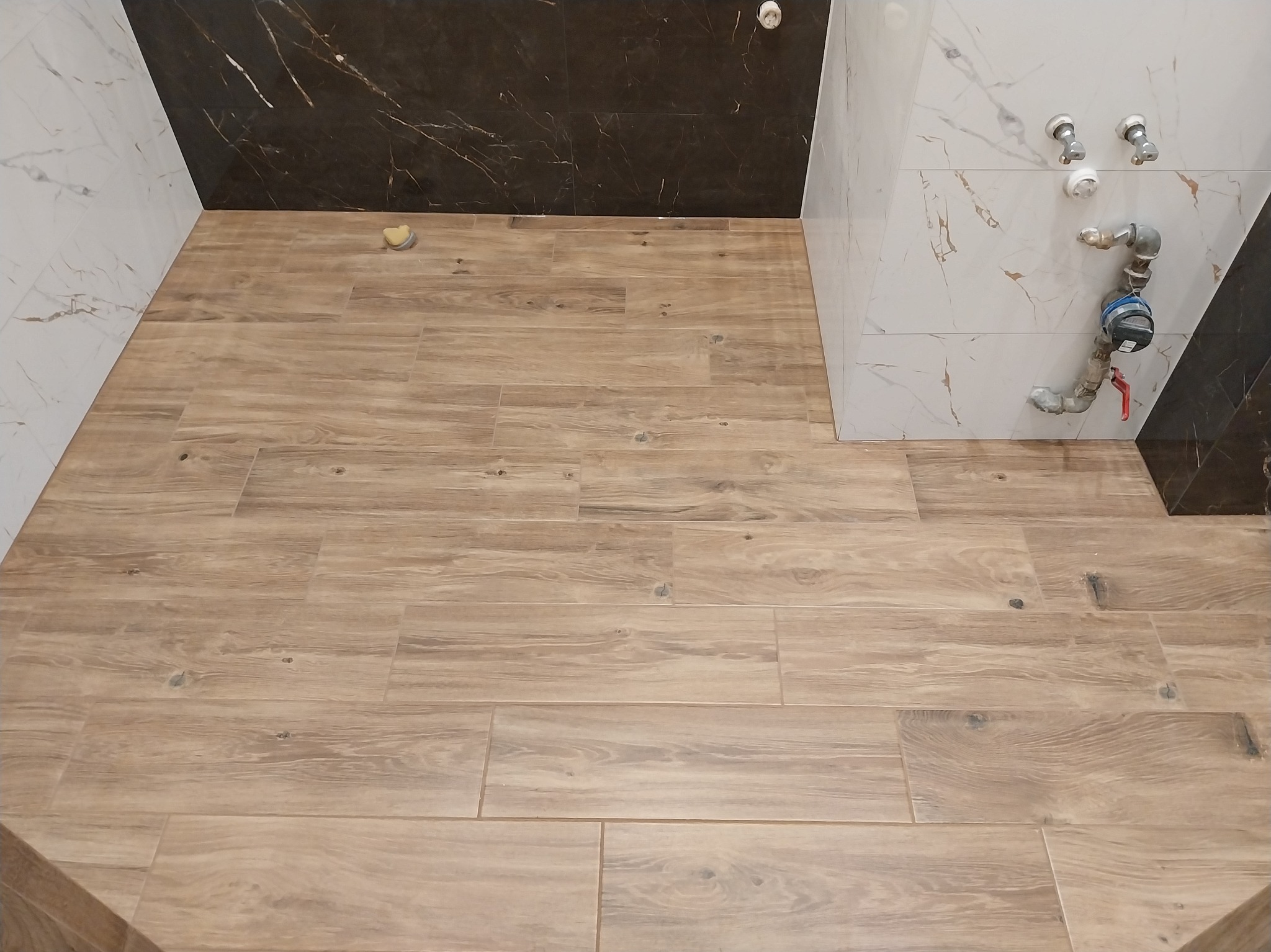 Ukończona podłoga w łazience wyłożona płytkami imitującymi drewno, z fragmentami ścian wykończonych marmurkowymi płytkami w odcieniach bieli i brązu.