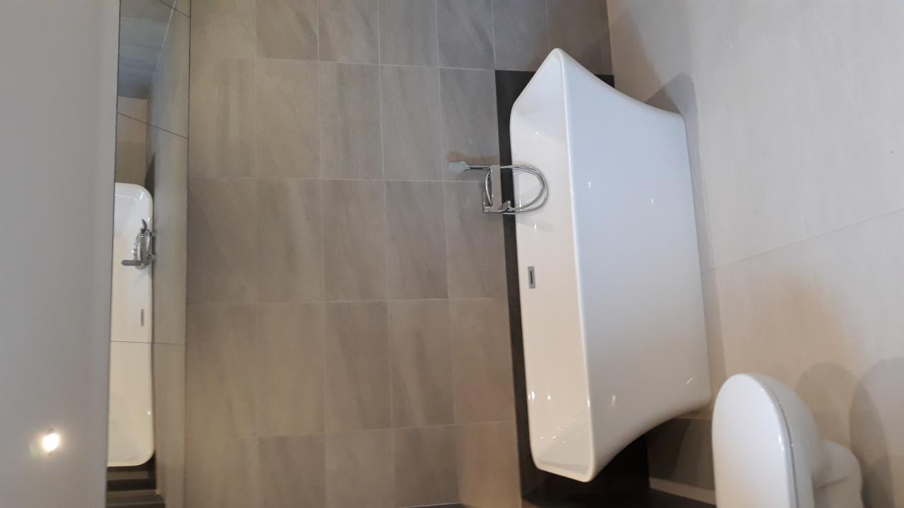 Nowoczesna łazienka z szarymi płytkami, białym pisuarze i toalecie, widoczna w lustrze, z chromowanym uchwytem na papier toaletowy.