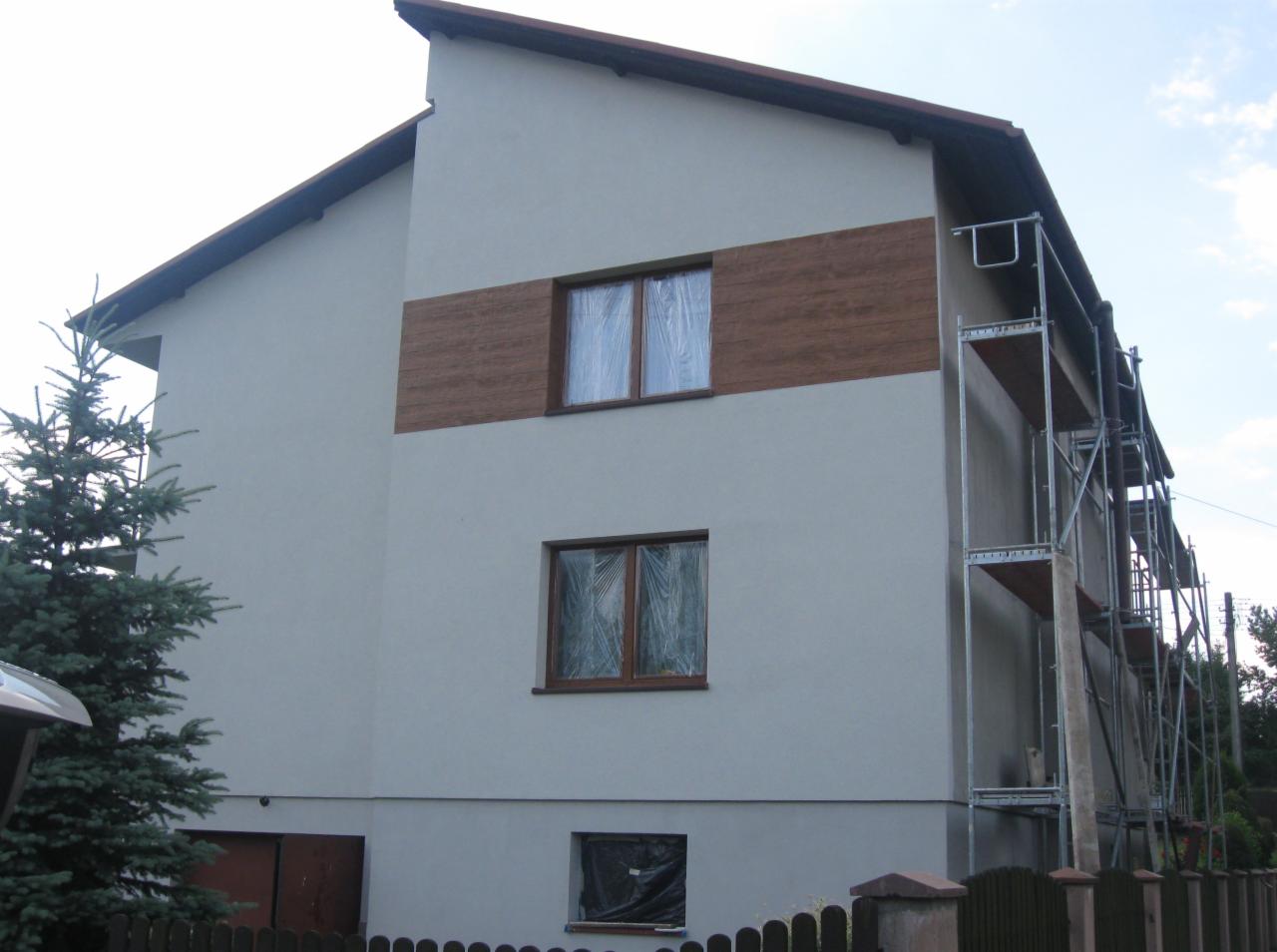 Dwukondygnacyjny dom po ociepleniu, z drewnianą wstawką na elewacji, okna zabezpieczone folią, rusztowanie z boku budynku, ogrodzenie i drzewo na pierwszym planie.