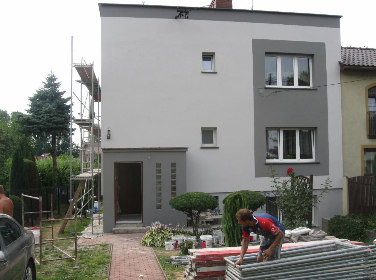 Dwukolorowa elewacja domu jednorodzinnego z pracownikiem przy rusztowaniu i przygotowanymi farbami. Widoczne detale architektoniczne i elementy wykończenia.