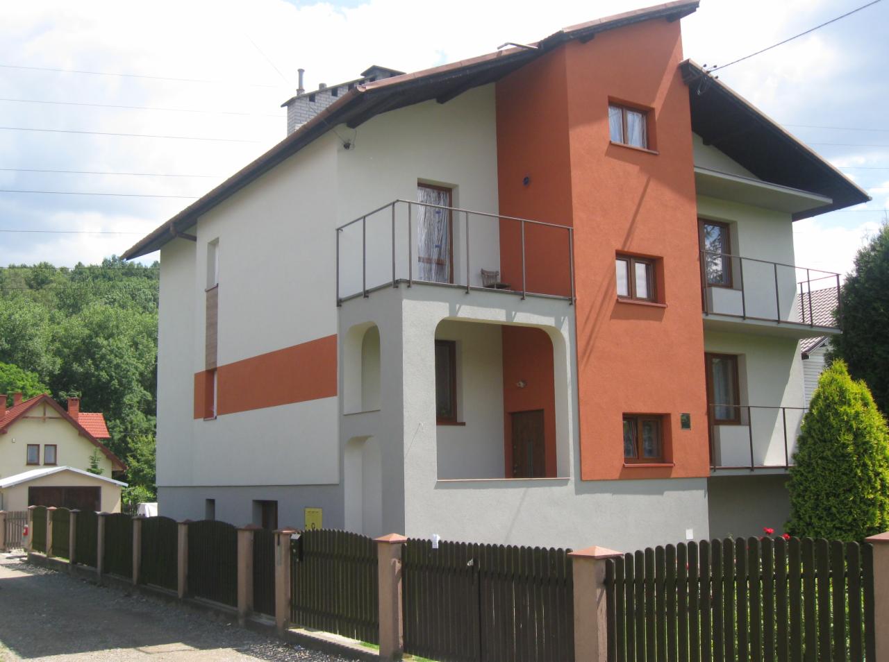 Dwukondygnacyjny dom z balkonami, pomalowany w odcieniach bieli, szarości i pomarańczy. Widoczna drewniana, pomalowana na ciemno zieleń brama i fragment zieleni.