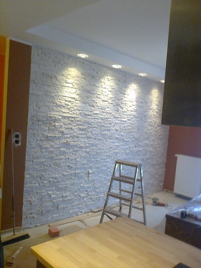 Ściana wyłożona dekoracyjnym kamieniem, oświetlona punktowymi lampami sufitowymi, z rozstawioną drabinką w trakcie prac wykończeniowych.