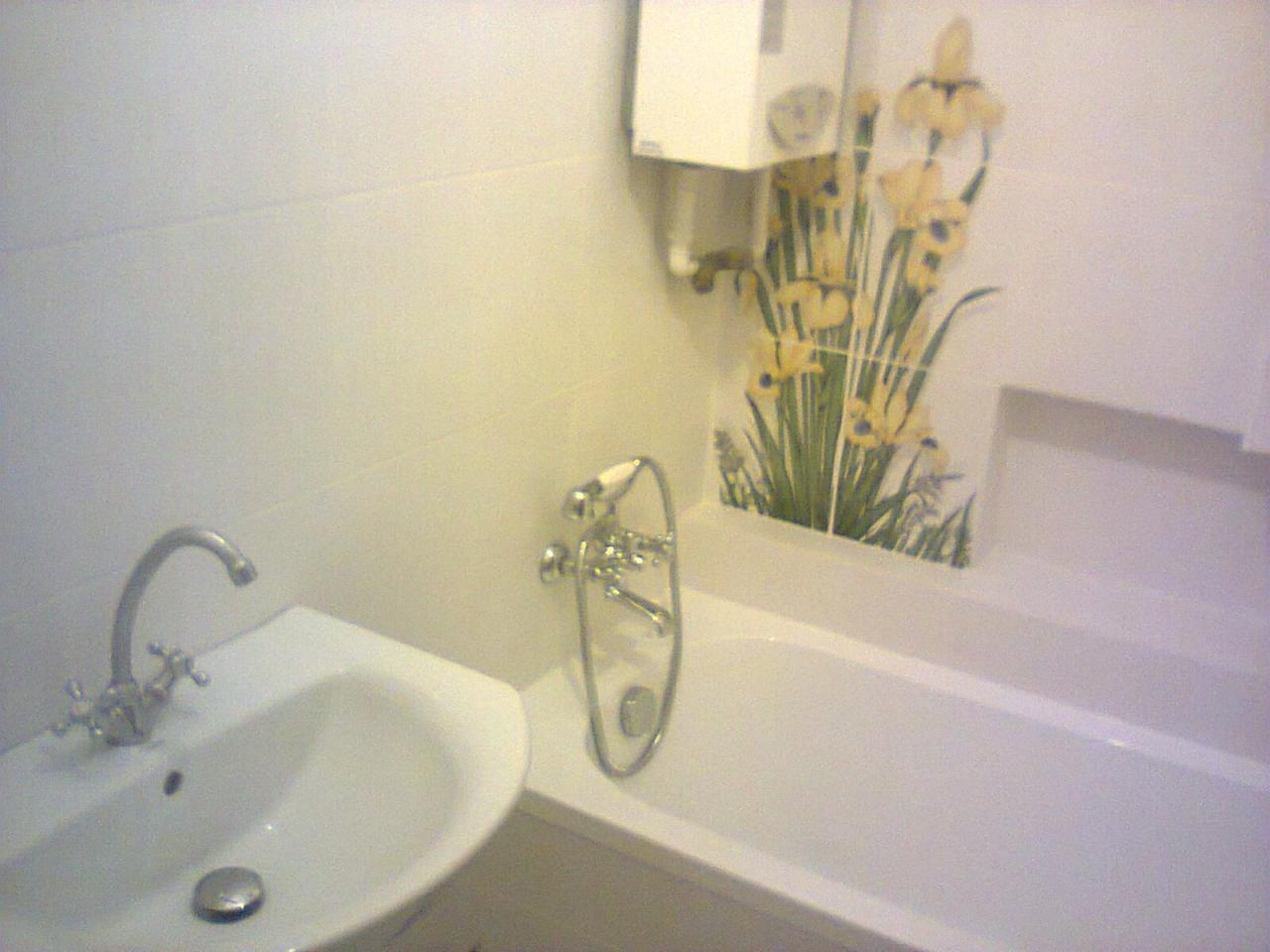 Biała łazienka z umywalką, wanną i baterią, ściany wyłożone jasnymi płytkami z dekoracją kwiatową, piec gazowy na ścianie.