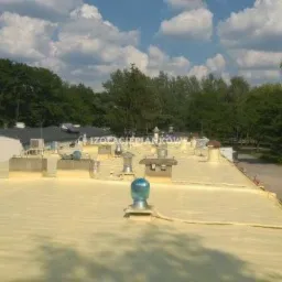 Żółta, gładka powierzchnia dachu pokryta pianką izolacyjną, widoczne kominy i elementy wentylacyjne, w tle budynki i drzewa pod błękitnym niebem z chmurami.